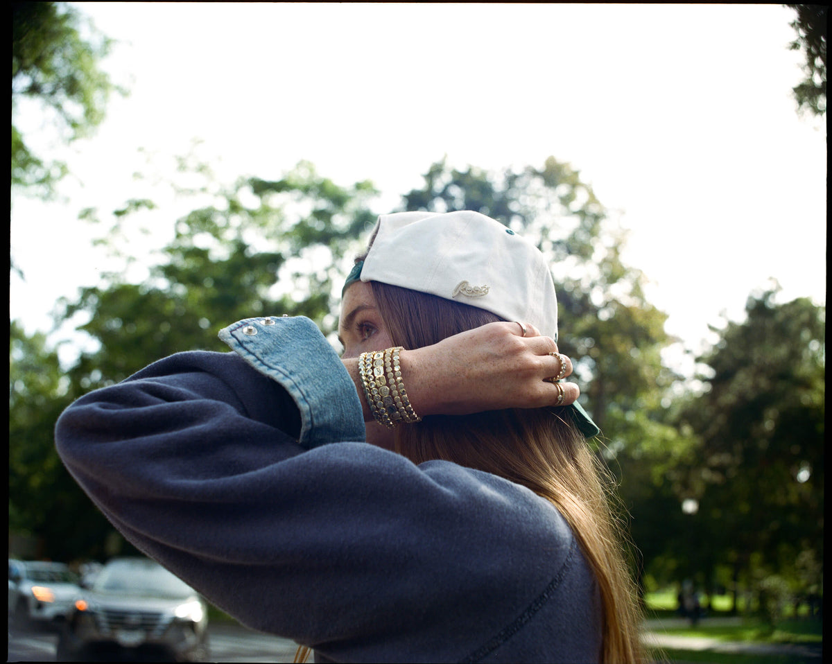 A person outdoors wearing a white baseball cap backwards, a dark blue sweatshirt, and multiple gold bracelets, with trees and a blurred street background.