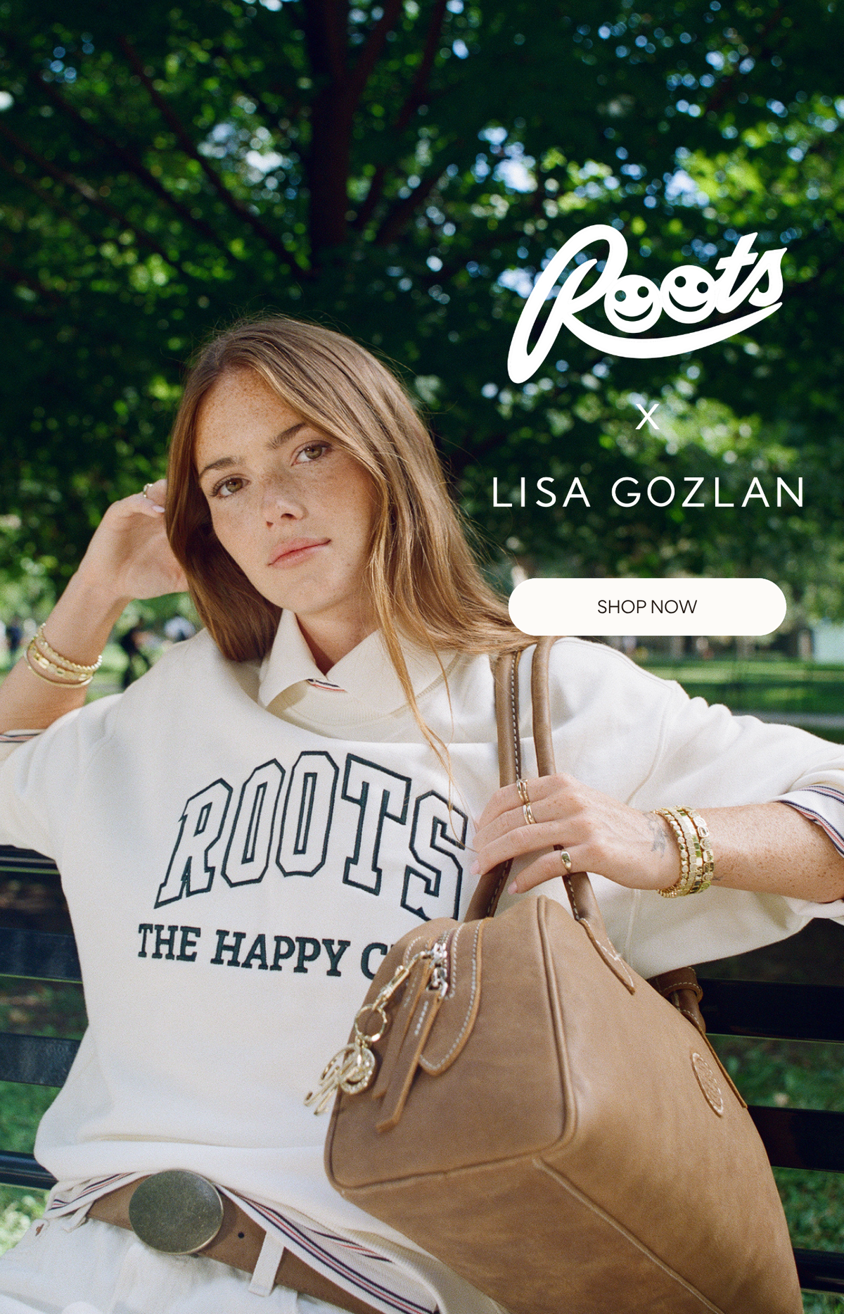 A woman sitting outdoors on a bench in a park-like setting, wearing a white Roots sweatshirt and holding a tan leather handbag. The image includes the Roots x Lisa Gozlan branding and a 'SHOP NOW' button.