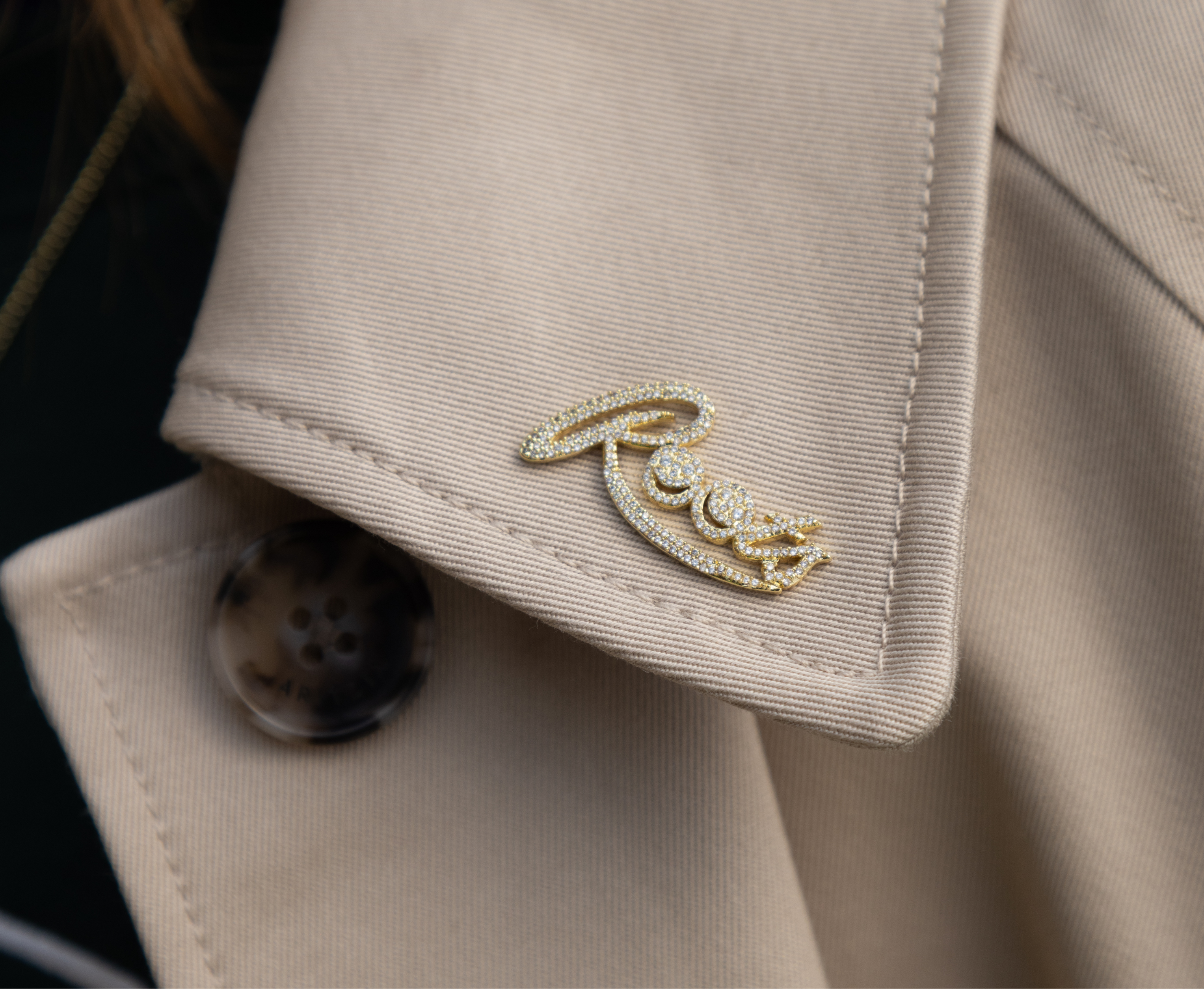 A close-up of a beige trench coat lapel featuring a gold-tone script pin with small clear stones spelling 'Rory', next to a brown tortoiseshell button.
